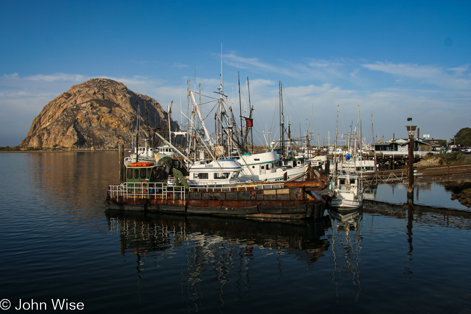 Early morning at Morro Bay, California with fishing boats in the foreground and Morro Rock in the background against a bright blue sky with a light band of clouds