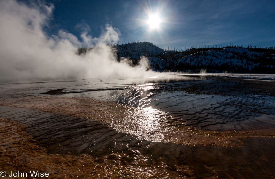 Grand Prismatic Spring with reflective water foreground and sun overhead on winter day at Midway Geyser Basin in Yellowstone National Park January 2010