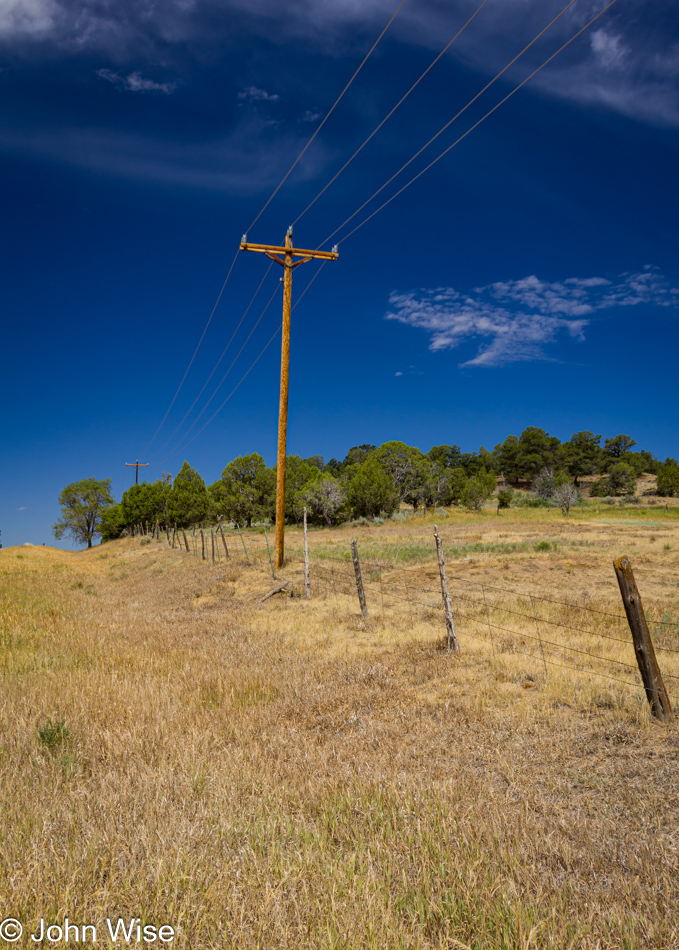 A telephone pole next to barbed wire fence in the dry grass with a deep blue sky